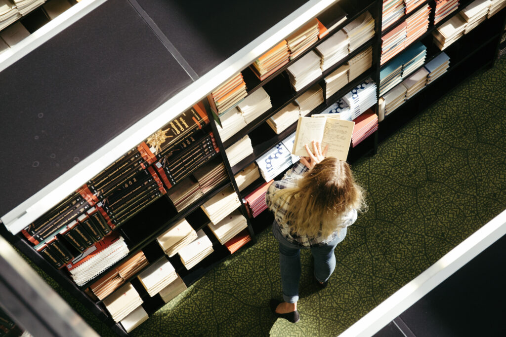 A young woman browsing bookshelves in a cozy library, reaching for a book with a focused expression, surrounded by warm lighting and rows of neatly arranged books.